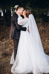 A newlywed couple poses in a forest, embracing warmly. The bride wears an elegant white dress and holds a bouquet of white flowers while the groom smiles beside her. A tender moment captured in nature