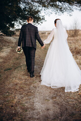 The newlywed couple walks along a natural path, holding hands. The bride in a long white dress looks back with a smile while the groom walks beside her. A warm and peaceful outdoor moment.