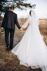 The newlywed couple walks along a natural path, holding hands. The bride in a long white dress looks back with a smile while the groom walks beside her. A warm and peaceful outdoor moment.