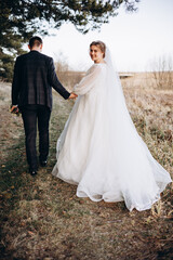 The newlywed couple walks along a natural path, holding hands. The bride in a long white dress looks back with a smile while the groom walks beside her. A warm and peaceful outdoor moment.