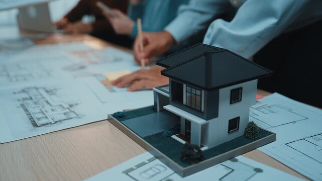 A diverse team of architects engages in a collaborative office meeting, reviewing detailed architectural plans and a 3D model of a house to inspire innovative project development. SACTR