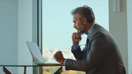Mature businessman in a suit concentrating on a laptop at a glass table in a modern office with...