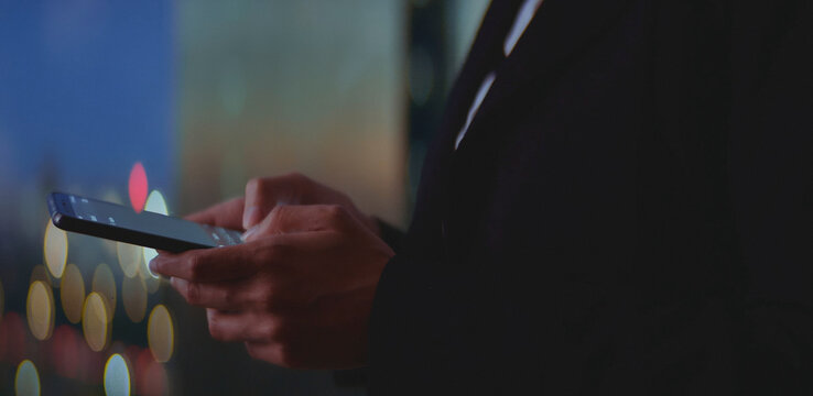 Business person using a smartphone, connecting to technology and communicating online while standing in an urban environment with bokeh city lights illuminating the dark night