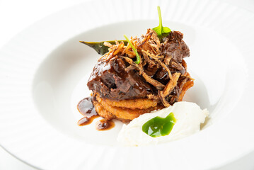 Braised beef short rib served over a base of golden-brown polenta cakes, topped with crispy fried onions and a rich brown sauce on a white plate.