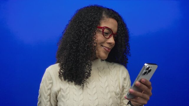 Woman wearing red glasses and cable knit sweater smiling while holding smartphone in studio; engagement.