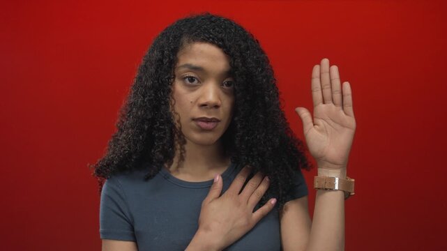 Woman with serious expression holding hand on chest and the other hand raised against red studio backdrop; sincerity.