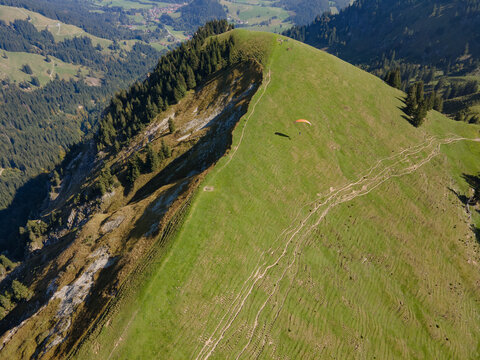 Aerial view of a paraglider soaring near the summit of a grassy mountain ridge, contrasting with the rugged, rocky slopes below, Bad Hindelang, Bayern, Deutschland.