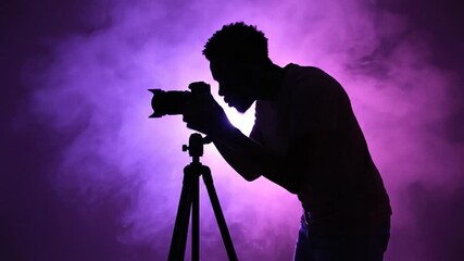 A silhouette of a photographer working on a tripod setup, surrounded by smoky purple light