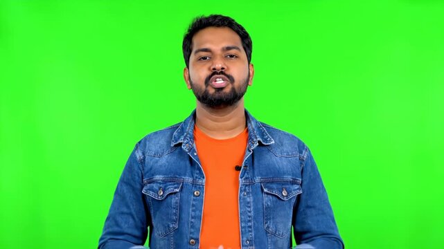 A young indian man with a beard, wearing a denim jacket and orange tshirt, speaking directly to the camera with an open mouth against a green screen vector illustration