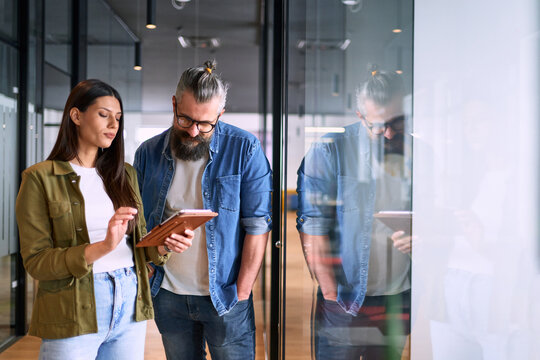 Two business professionals having a focused discussion while reviewing information on a tablet in a bright modern office hallway, symbolizing teamwork, communication, and digital collaboration.