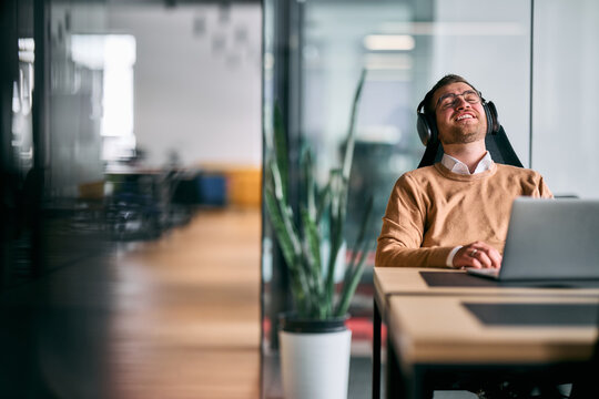 Happy businessman sitting at office desk, wearing headphones and relaxing while listening to music during a break, representing stress relief, balance, and positive work atmosphere in modern office - Powered by Adobe