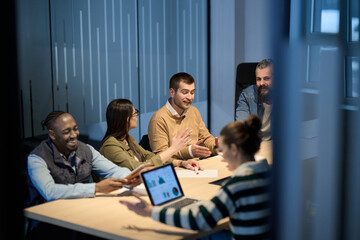 Diverse business team gathered around a table discussing analytics during a meeting in a modern office. Laptop screen shows charts and graphs, symbolizing teamwork, communication, and data driven