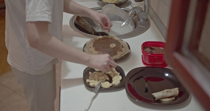 Woman in pajamas prepares a sweet breakfast of crepes with chocolate spread and sliced banana in home kitchen.