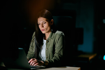 Concentrated businesswoman working late at night on laptop in dimly lit office, representing...