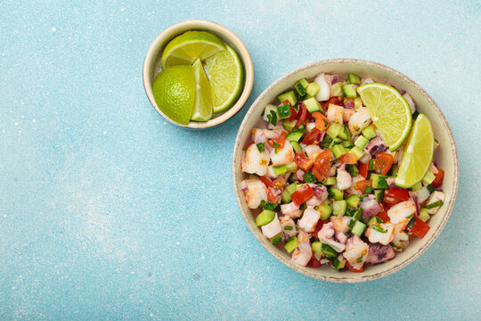Fresh and vibrant peruvian seafood ceviche with shrimps, octopus, tomatoes, cucumber and parsley, served with lime in a bowl on a light blue background from above, dish of Peru. Copy space