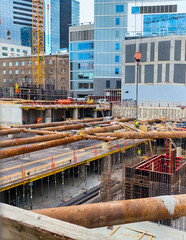 A view of a large, deep construction pit in an urban area, showing multiple levels of scaffolding, concrete forms, steel rebar, and large rusty support pipes, surrounded by modern glass skyscrapers. 