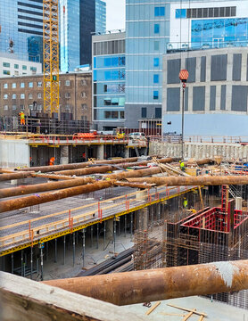 A view of a large, deep construction pit in an urban area, showing multiple levels of scaffolding, concrete forms, steel rebar, and large rusty support pipes, surrounded by modern glass skyscrapers. 