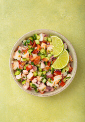 Fresh and vibrant peruvian seafood ceviche with shrimps, octopus, tomatoes, cucumber and parsley, served with lime in a bowl on a light green background from above, dish of Peru