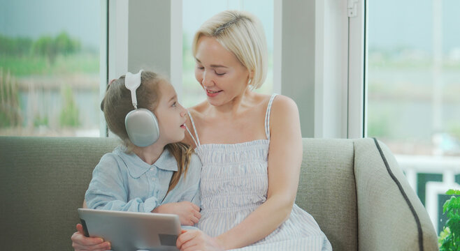 Caucasian mother and little daughter wearing headphones listening to music with tablet at home, smiling and bonding through fun and joyful family time.