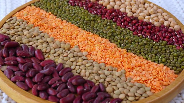 Colorful selection of raw legumes, including beans, lentils, and chickpeas, arranged in neat rows on a wooden plate, slowly rotating. Healthy food ingredients rich in protein and fiber