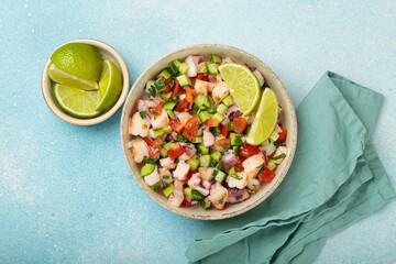 Fresh and vibrant peruvian seafood ceviche with shrimps, octopus, tomatoes, cucumber and parsley, served with lime in a bowl on a light blue background from above, dish of Peru