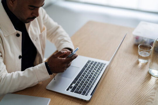 Side view of serious Arab male remote worker messaging on cellphone while sitting at table with netbook in modern apartment during workday