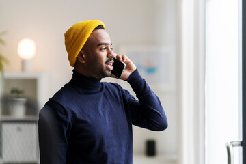 Bearded young Arab male in casual turtleneck and bright hat standing near window and having conversation via cellphone in apartment