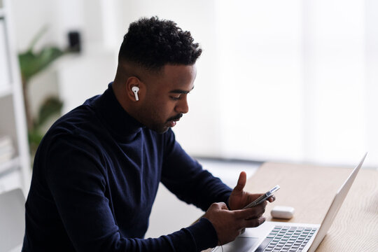 Arab male teleworker in true wireless earbuds surfing cellphone while working on netbook in home office and looking at screen with wide smile
