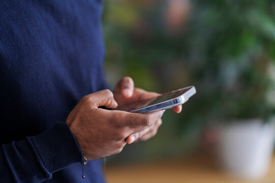 Crop anonymous Arab male in warm sweater messaging on cellphone while standing against blurred houseplants at soft daylight