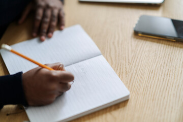 High angle of crop anonymous Arab male writing ideas in planner at wooden table with smartphone at daylight