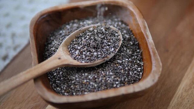 Close up top view of raw chia seeds slowly falling from a wooden spoon into a rustic bowl, highlighting a healthy diet, superfood nutrition, and organic natural ingredients for wellness