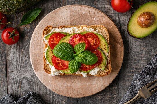Overhead shot of a creamy cheese sandwich garnished with fresh basil, red pepper slices, avocado half, and cherry tomatoes on fabric over rustic wooden surface
