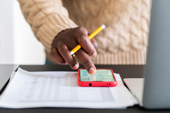 Crop anonymous black male in casual sweater with documents and pencil using smartphone counting bills on calculator