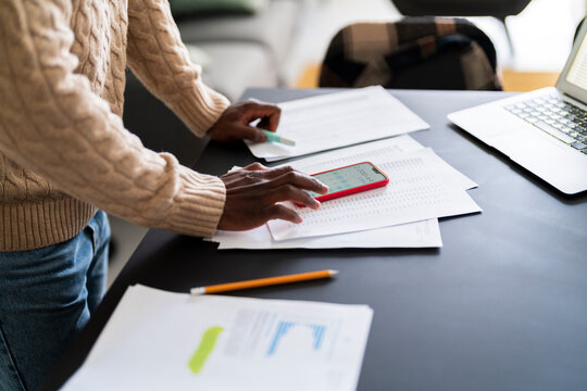 High angle of crop anonymous black male in casual outfit standing at table with papers laptop pencil using smartphone calculating bills at home