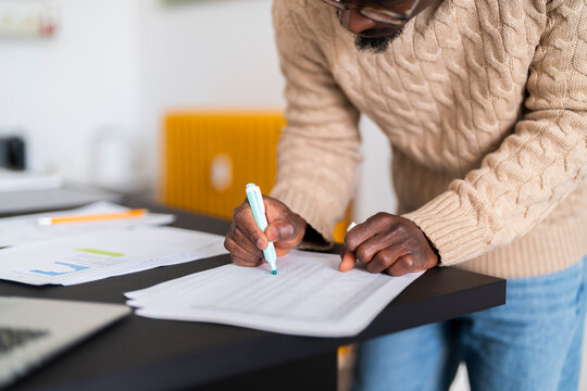 Crop black male in sweater and jeans leaning forward standing at table highlighting payment for apartment with green marker while checking bills