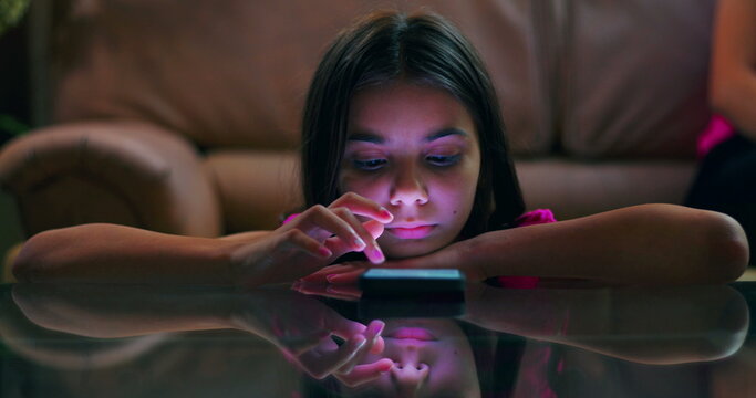 Young girl resting her head on arms while using smartphone on glass table, focused and engaged with screen in dim indoor setting, glowing light on face - Powered by Adobe
