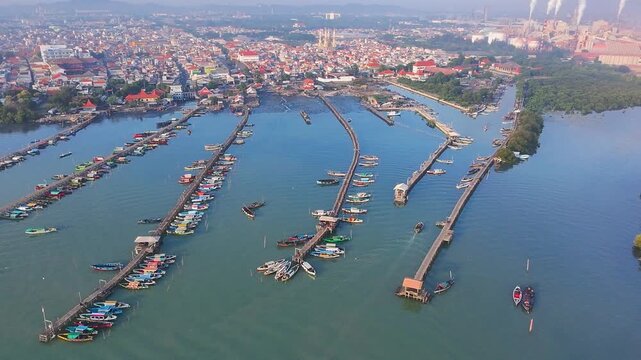 Aerial view of vibrant fishing boats docked at long wooden piers in a harbor near Lumpur, Gresik, East Java, with a dense coastal town skyline.
