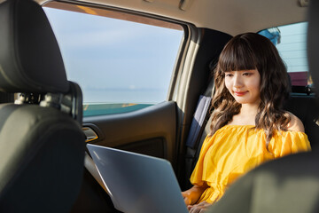 Joyful woman enjoying a carefree moment using laptop while sitting on a backseat of a car overlooking the sea and Clear blue sky with a few clouds during a sunny day, embodying freedom and happiness.