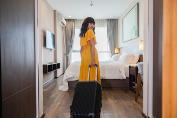 Friendly confident woman enters a hotel room, pulling suitcase, looking back at the camera and smiling. She looks ready to settle in hotel. Hotel room interior and Cozy bedroom setting in background.