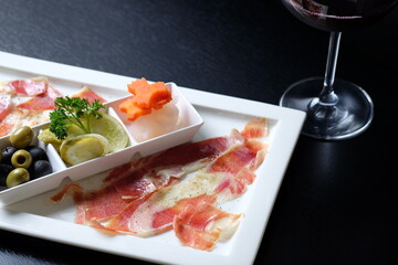 An elegant platter of assorted food with a glass of red wine. Featuring olives, pickles, and prosciutto arranged on a sleek, minimalist surface, creating an appetizing visual feast