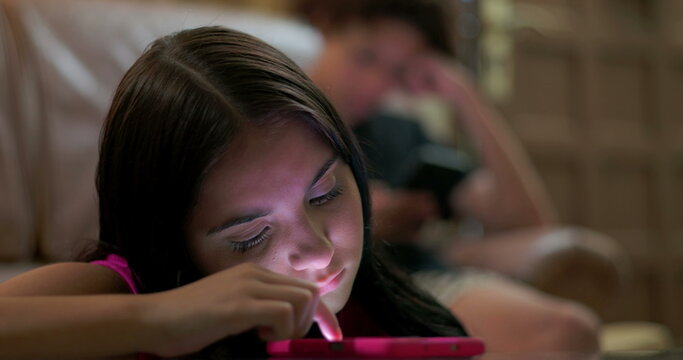 Girl intensely scrolling on smartphone with glowing light reflecting on face, boy blurred in background on sofa also using phone, highlighting the impact of digital overuse