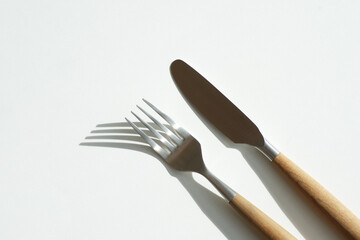  Knife and fork with wooden handle lie on white table. The fork casts an elegant shadow,  top view.