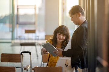 Relaxed asian couple casually sitting in airport with suitcase, using travel application, travel booking, online promotion, checking flight information, playing social media on tablet at airport.  