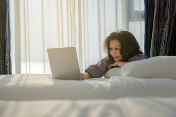 Relaxed asian middle aged woman sitting on comfortable bed at home or hotel and using laptop looking at screen working in internet, enjoying a new day, feeling rested and refreshed. Copy space