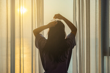 Young woman styling hair with a claw clip ,easy daily hairstyling. Woman standing at window in bedroom and fresh wake up with sunrise in hotel or home ready for day. View and nature on holiday. 