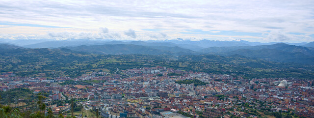 Expansive panoramic vista of urban settlement surrounded by layered mountain ranges and rolling hills beneath moody sky