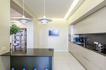 A modern kitchen featuring sleek cabinets, a dark countertop, and stylish pendant lights. A TV is mounted on the wall