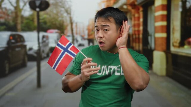 Young asian man holding a norwegian flag on a city street, playfully making a listening gesture with his hand to his ear, giving a sense of attention and curiosity.