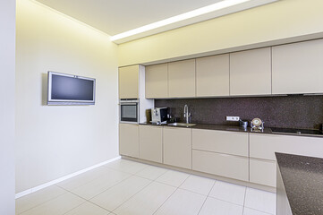 A modern kitchen with sleek beige cabinets, dark countertops, and a TV on the wall. The floor is light-colored tile