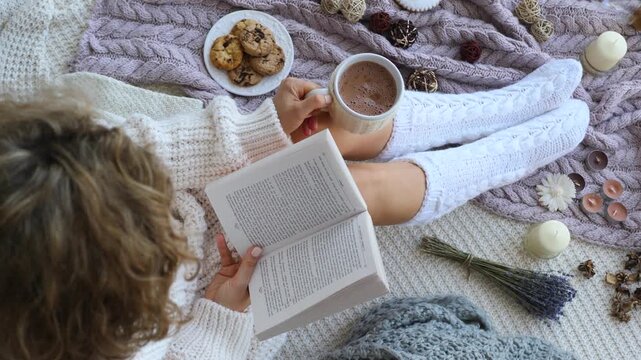 Cozy scene woman in knitted sweater and warm knee socks reads book on blanket. Female holds hot cocoa while immersing herself in book. Book captures attention, creating atmosphere calm and comfort.
